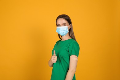 Vaccinated woman with protective mask and medical plaster on her arm showing thumb up against yellow background Photo of Vaccinated woman with protective mask and medical plaster on her arm showing thumb up against yellow background