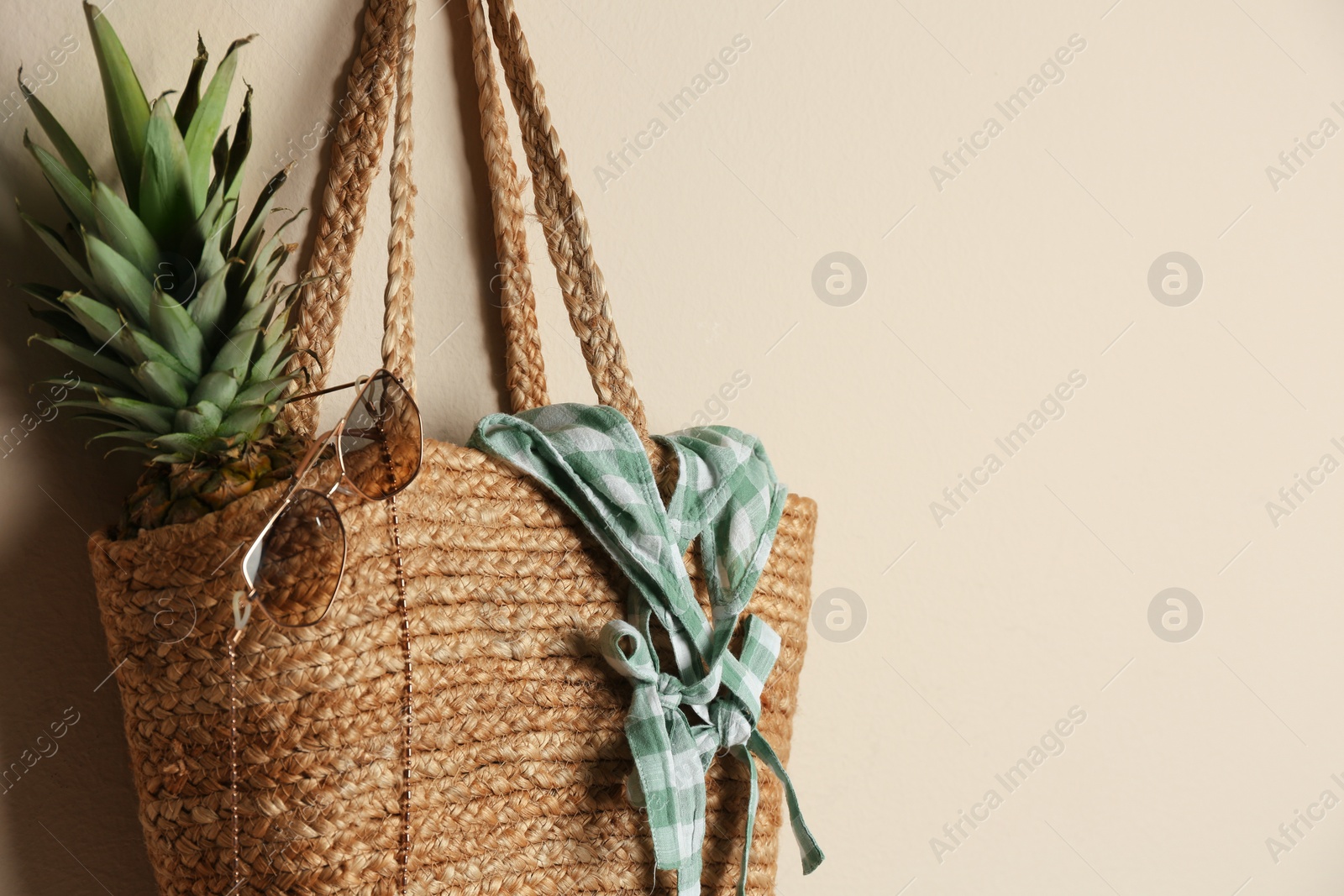 Elegant woman's straw bag with pineapple and accessories hanging on beige background. Space for text Photo of Elegant woman's straw bag with pineapple and accessories hanging on beige background. Space for text