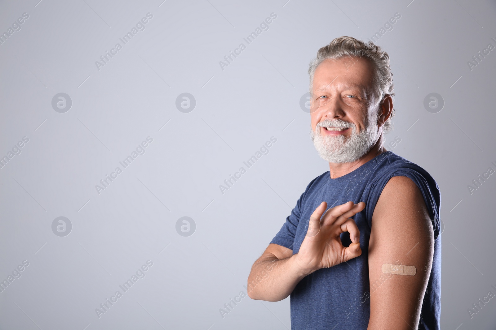 Cheerful senior man showing arm with bandage after vaccination on grey background. Space for text Photo of Cheerful senior man showing arm with bandage after vaccination on grey background. Space for text