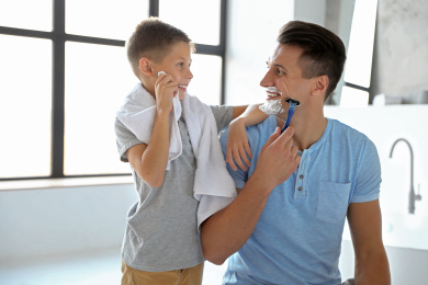 Son wiping face with towel while his dad shaving in bathroom Photo of Son wiping face with towel while his dad shaving in bathroom