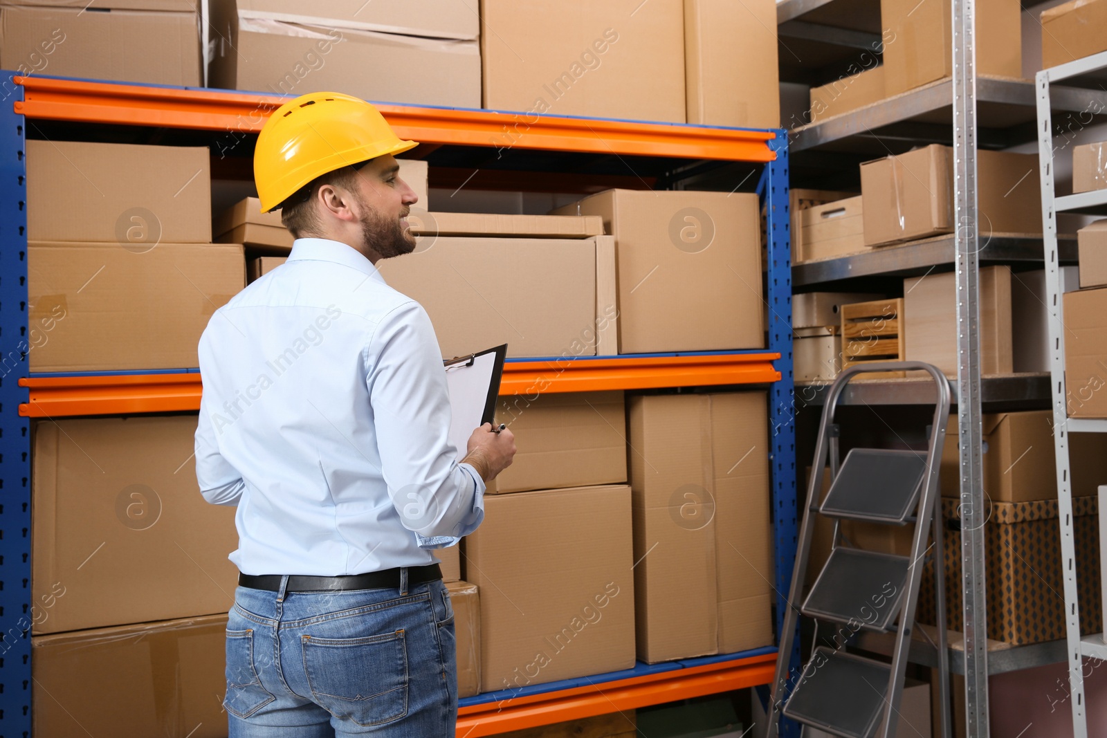 Young man with clipboard near rack of cardboard boxes at warehouse, back view Photo of Young man with clipboard near rack of cardboard boxes at warehouse, back view
