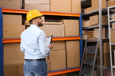 Young man with clipboard near rack of cardboard boxes at warehouse, back view Photo of Young man with clipboard near rack of cardboard boxes at warehouse, back view