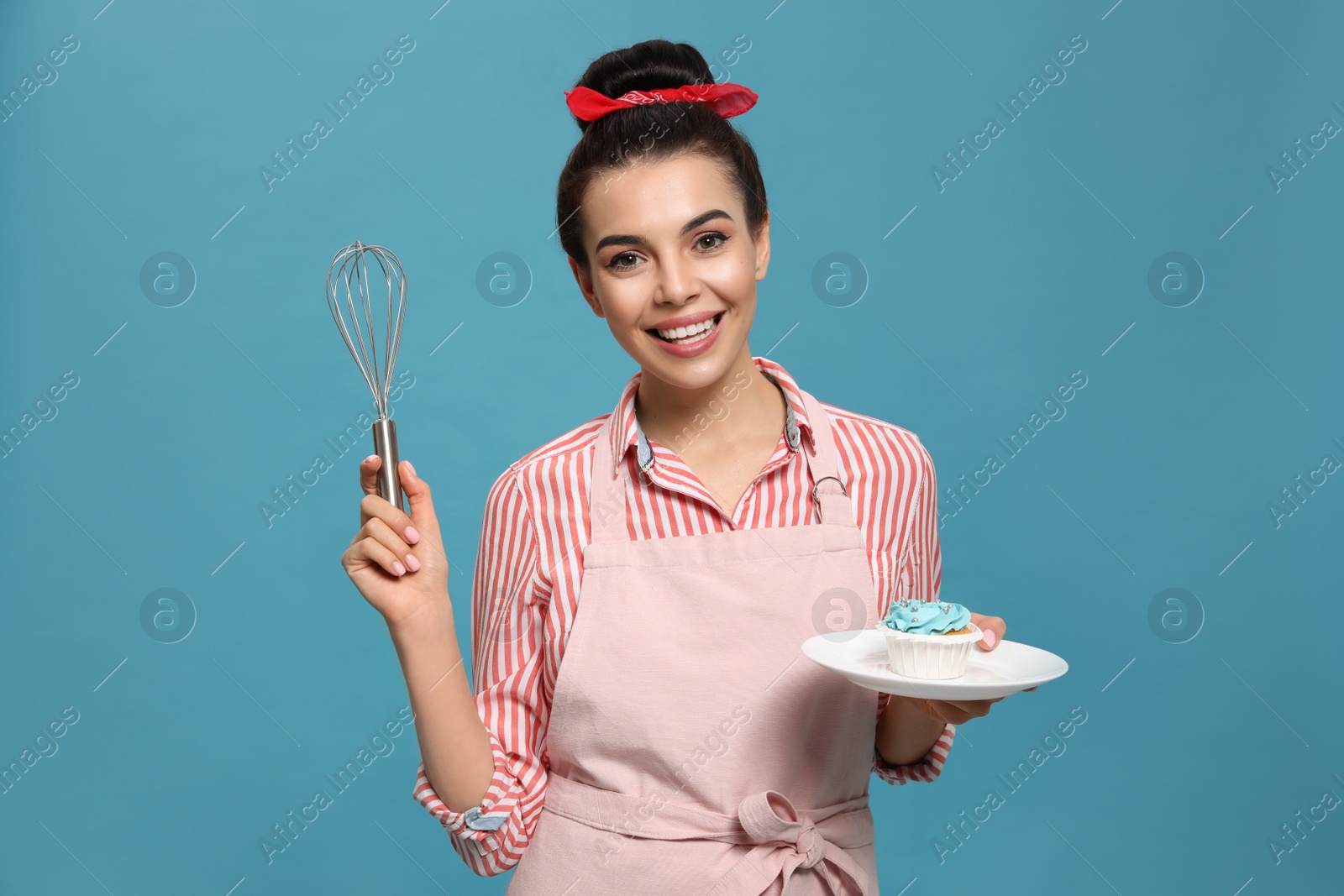 Young housewife with tasty cupcake and whisk on light blue background Photo of Young housewife with tasty cupcake and whisk on light blue background