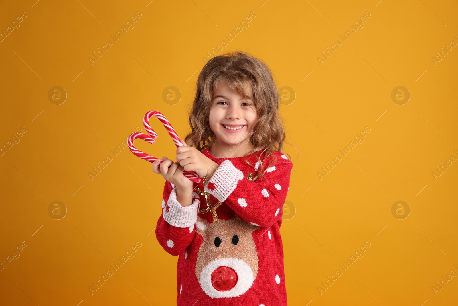 Cute little girl in Christmas sweater making heart shape with candy canes against orange background Photo of Cute little girl in Christmas sweater making heart shape with candy canes against orange background