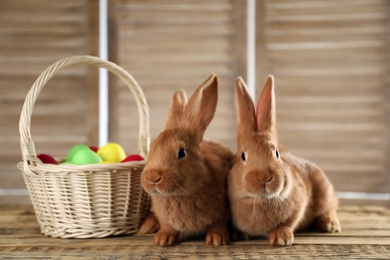 Cute bunnies and basket with Easter eggs on wooden table against blurred background Photo of Cute bunnies and basket with Easter eggs on wooden table against blurred background