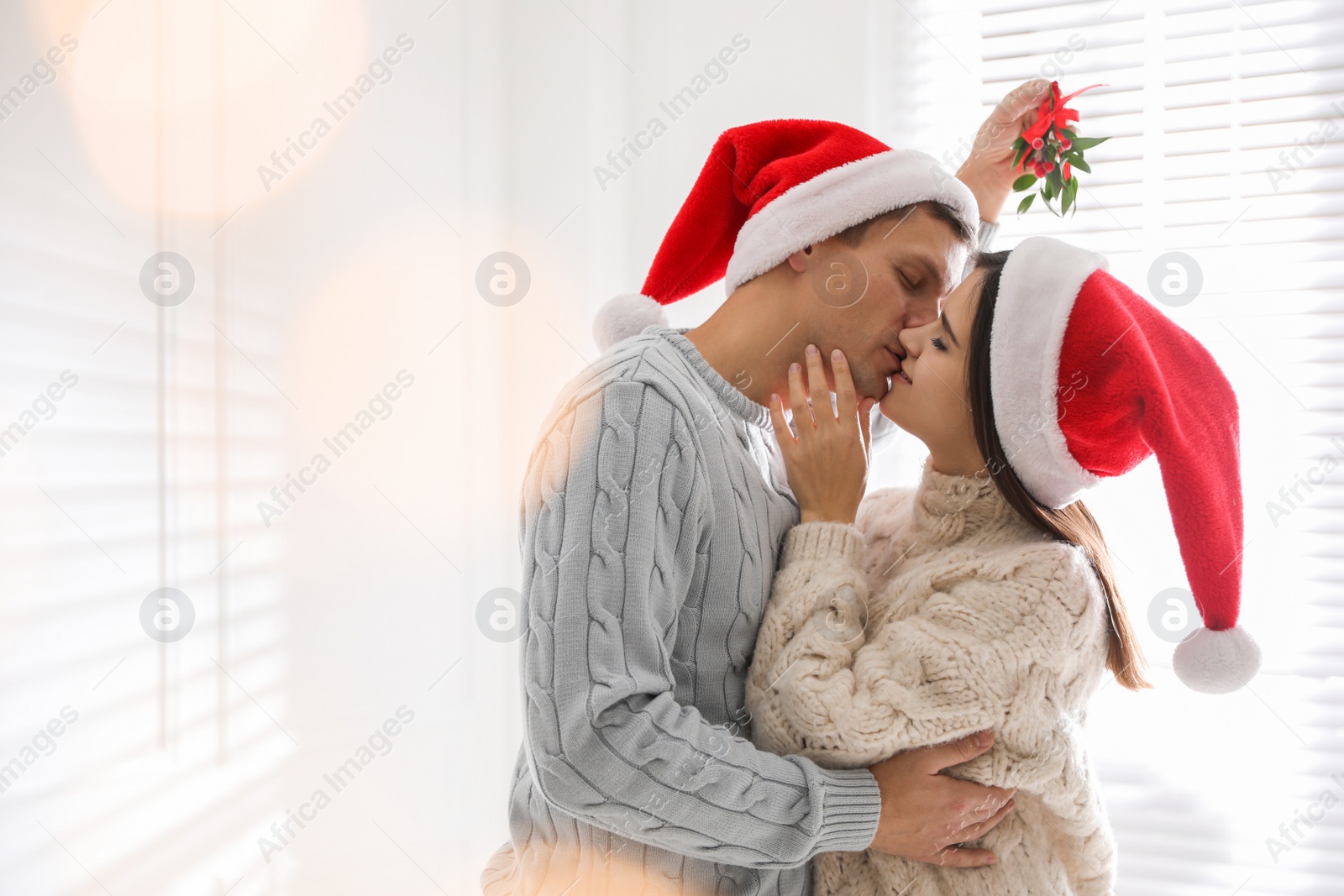 Happy couple in Santa hats kissing under mistletoe bunch at home, bokeh effect. Space for text Photo of Happy couple in Santa hats kissing under mistletoe bunch at home, bokeh effect. Space for text