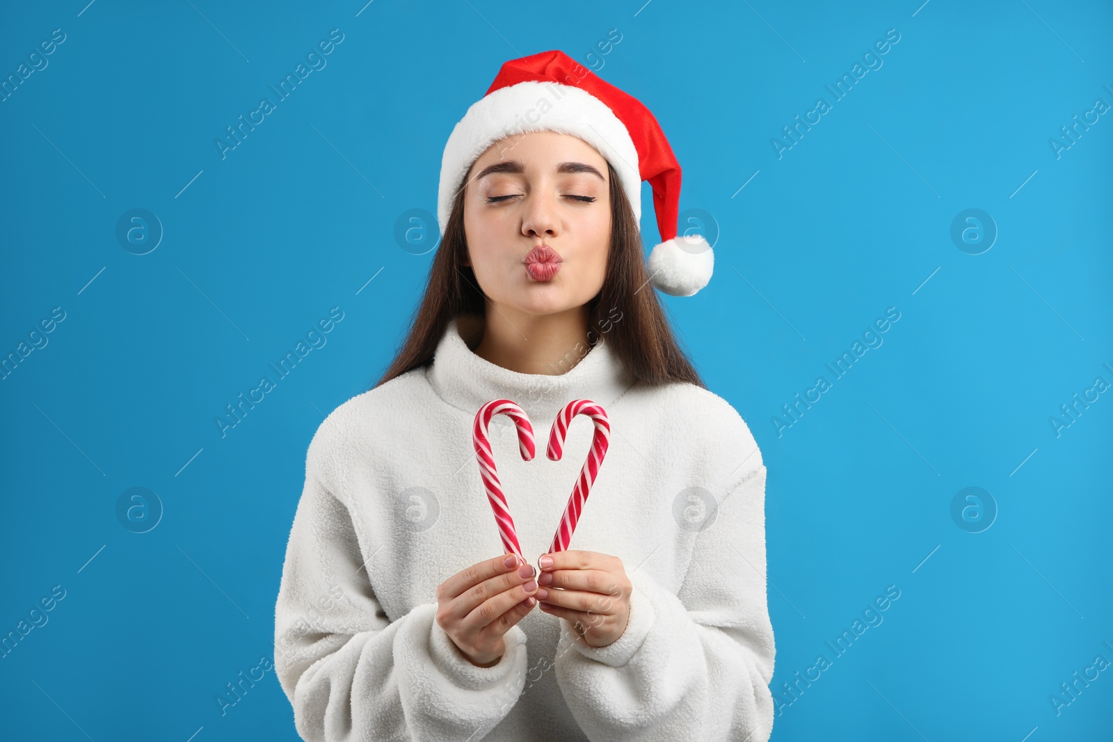 Young woman in beige sweater and Santa hat holding candy canes on blue background. Celebrating Christmas Photo of Young woman in beige sweater and Santa hat holding candy canes on blue background. Celebrating Christmas