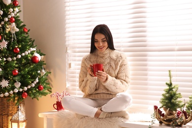 Young woman with cup of hot drink near Christmas tree at home Photo of Young woman with cup of hot drink near Christmas tree at home