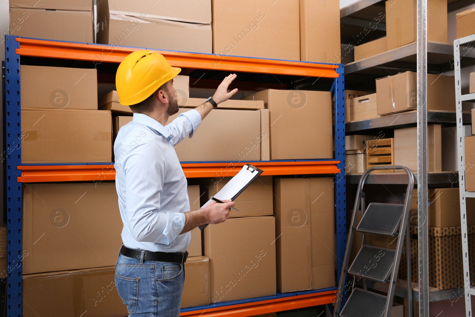 Young man with clipboard near rack of cardboard boxes at warehouse Photo of Young man with clipboard near rack of cardboard boxes at warehouse