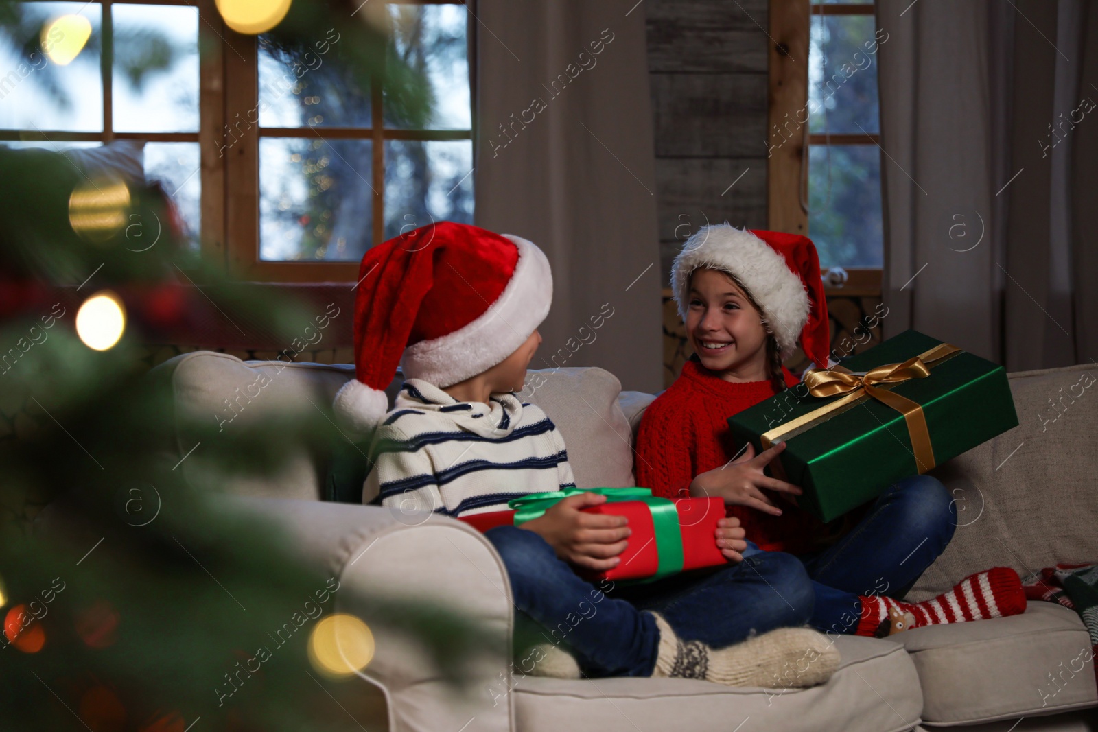 Happy children wearing Santa hats with Christmas gifts on sofa at home Photo of Happy children wearing Santa hats with Christmas gifts on sofa at home