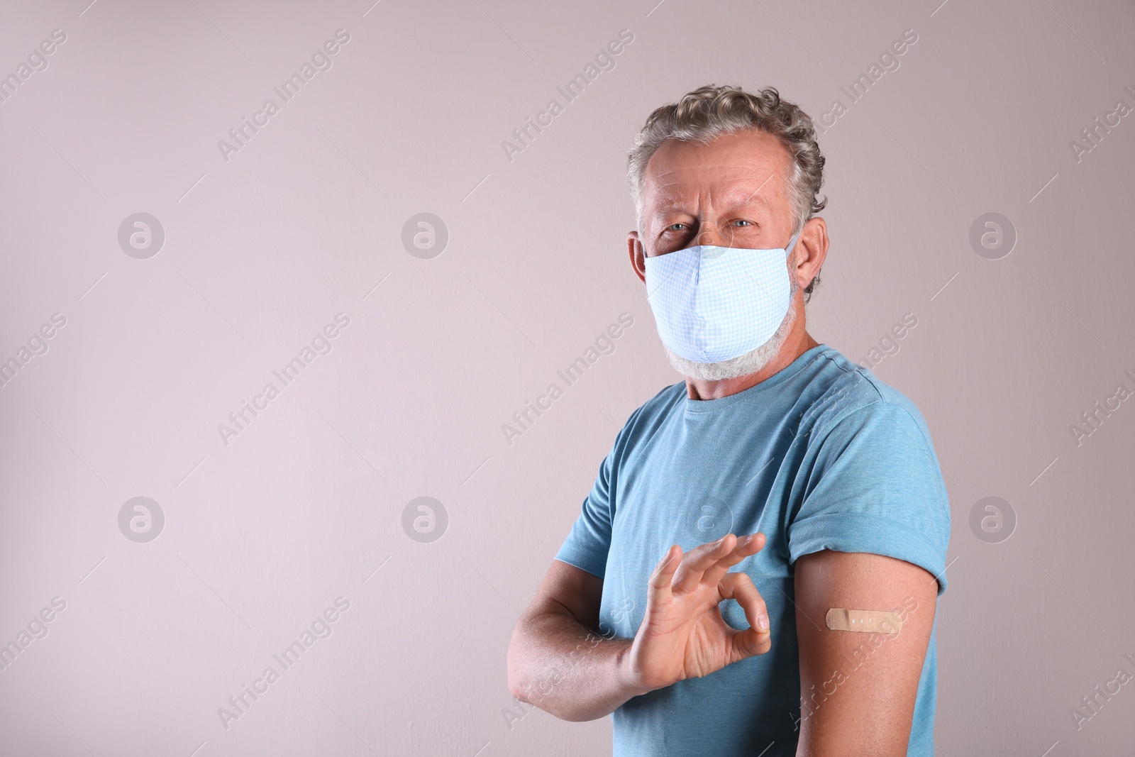 Cheerful senior man in protective mask showing arm with bandage after vaccination on beige background. Space for text Photo of Cheerful senior man in protective mask showing arm with bandage after vaccination on beige background. Space for text