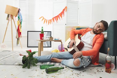 Young man with guitar sleeping near sofa in messy room after party Photo of Young man with guitar sleeping near sofa in messy room after party