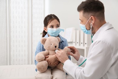 Pediatrician playing with little girl during visit in hospital. Doctor and patient wearing protective masks Photo of Pediatrician playing with little girl during visit in hospital. Doctor and patient wearing protective masks