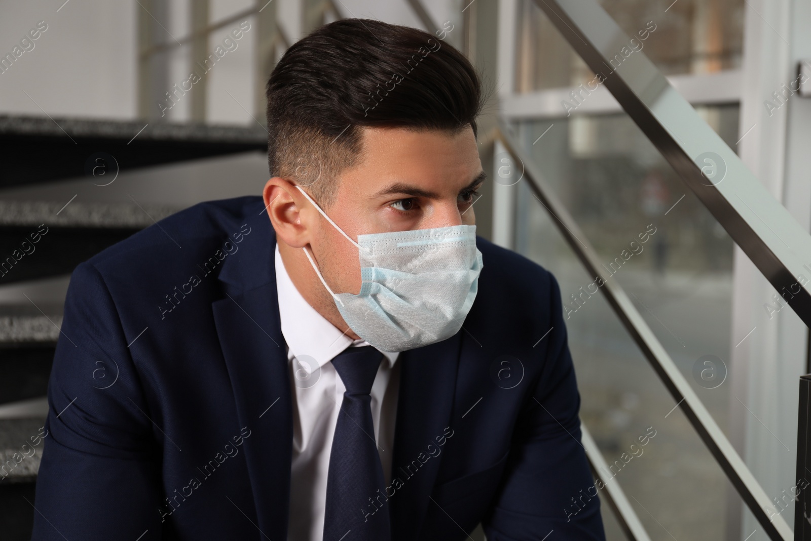 Sad man in protective mask on stairs indoors. Self-isolation during coronavirus pandemic Photo of Sad man in protective mask on stairs indoors. Self-isolation during coronavirus pandemic