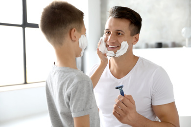 Son applying shaving foam onto father's face in bathroom Photo of Son applying shaving foam onto father's face in bathroom