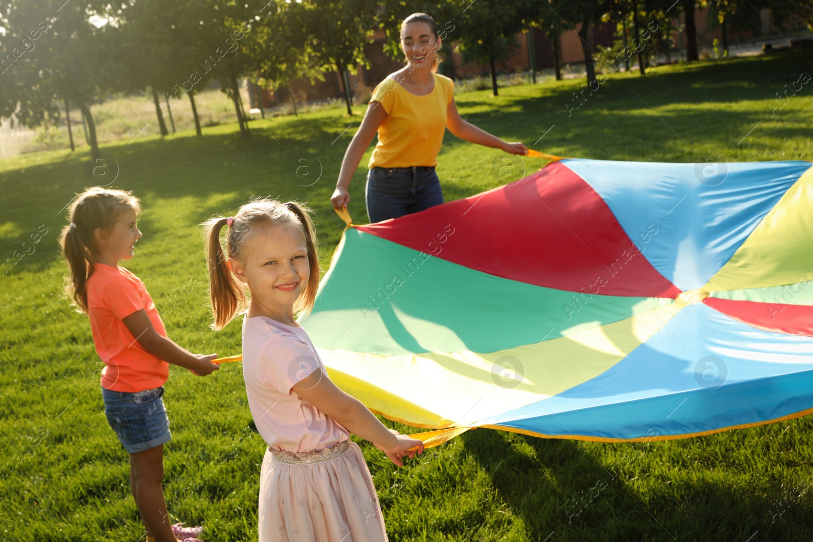 Photo of Group of children and teacher playing with rainbow playground parachute on green grass. Summer camp activity