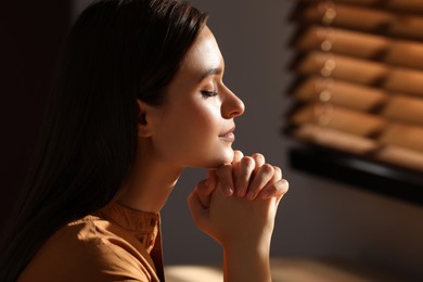 Religious young woman with clasped hands praying indoors Photo of Religious young woman with clasped hands praying indoors