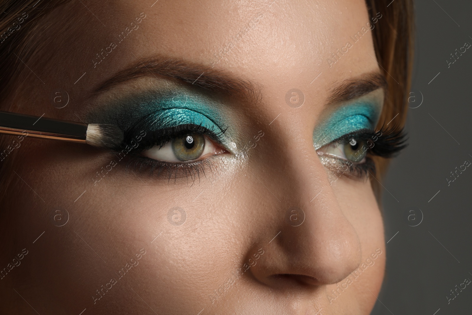 Applying eye shadow onto woman's face on grey background, closeup. Beautiful evening makeup Photo of Applying eye shadow onto woman's face on grey background, closeup. Beautiful evening makeup