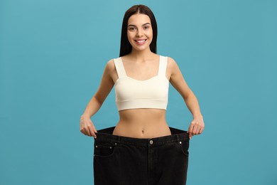 Happy young woman wearing big jeans after weight loss on turquoise background Photo of Happy young woman wearing big jeans after weight loss on turquoise background
