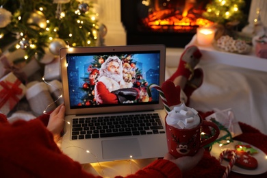 MYKOLAIV, UKRAINE - DECEMBER 23, 2020: Woman with sweet drink watching The Christmas Chronicles movie on laptop near fireplace at home, closeup. Cozy winter holidays atmosphere Photo of MYKOLAIV, UKRAINE - DECEMBER 23, 2020: Woman with sweet drink watching The Christmas Chronicles movie on laptop near fireplace at home, closeup. Cozy winter holidays atmosphere