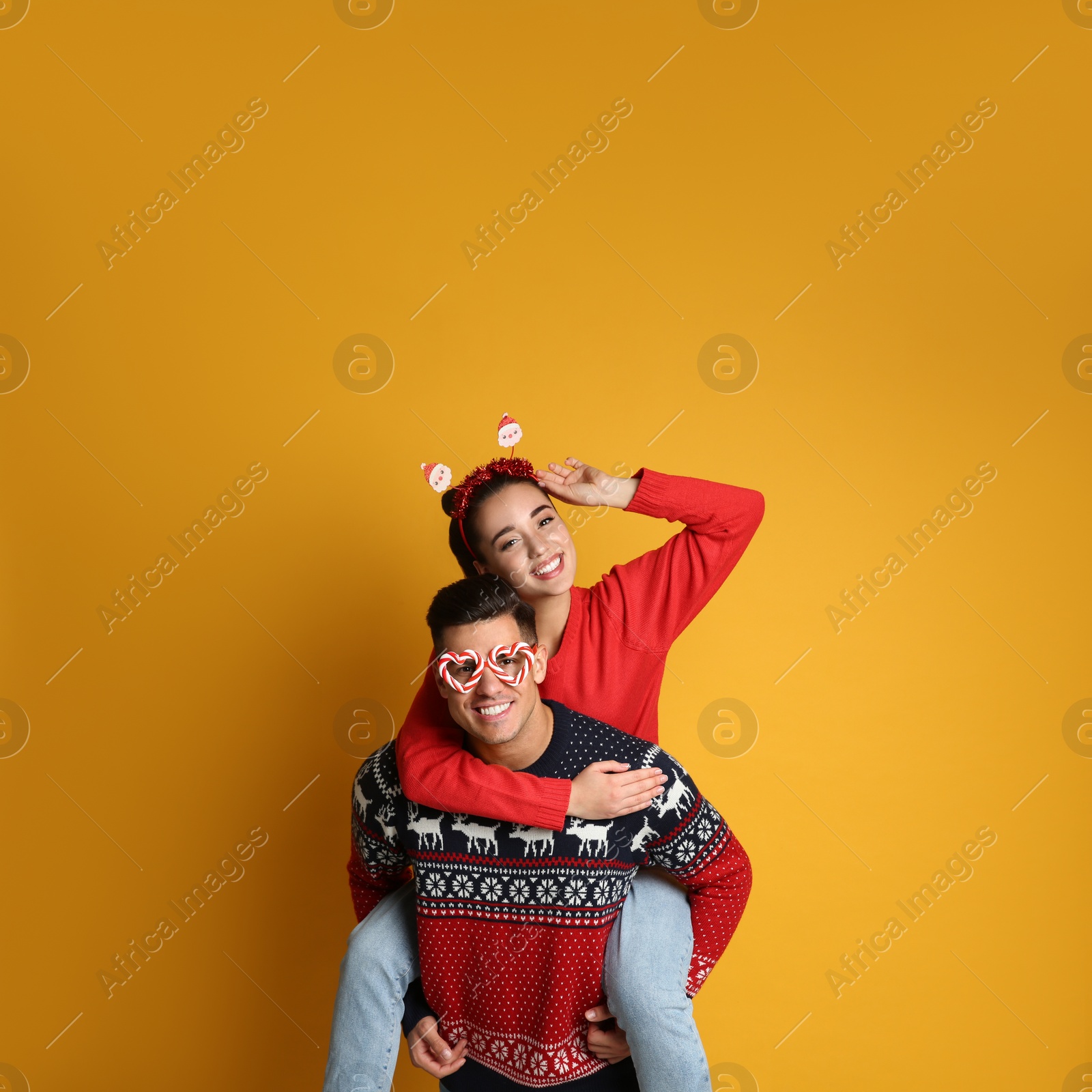 Couple in Christmas sweaters, Santa headband and party glasses on yellow background Photo of Couple in Christmas sweaters, Santa headband and party glasses on yellow background