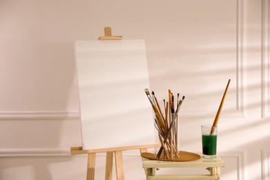 Wooden easel with empty canvas and brushes in art studio Photo of Wooden easel with empty canvas and brushes in art studio