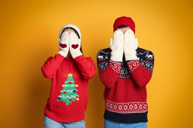 Couple in Christmas sweaters, knitted mittens and hats hiding faces on yellow background Photo of Couple in Christmas sweaters, knitted mittens and hats hiding faces on yellow background