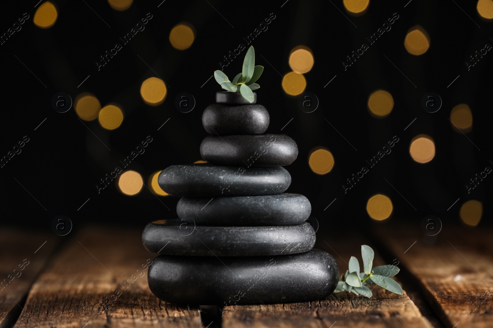 Stack of black spa stones with green branches on wooden table against blurred lights Photo of Stack of black spa stones with green branches on wooden table against blurred lights