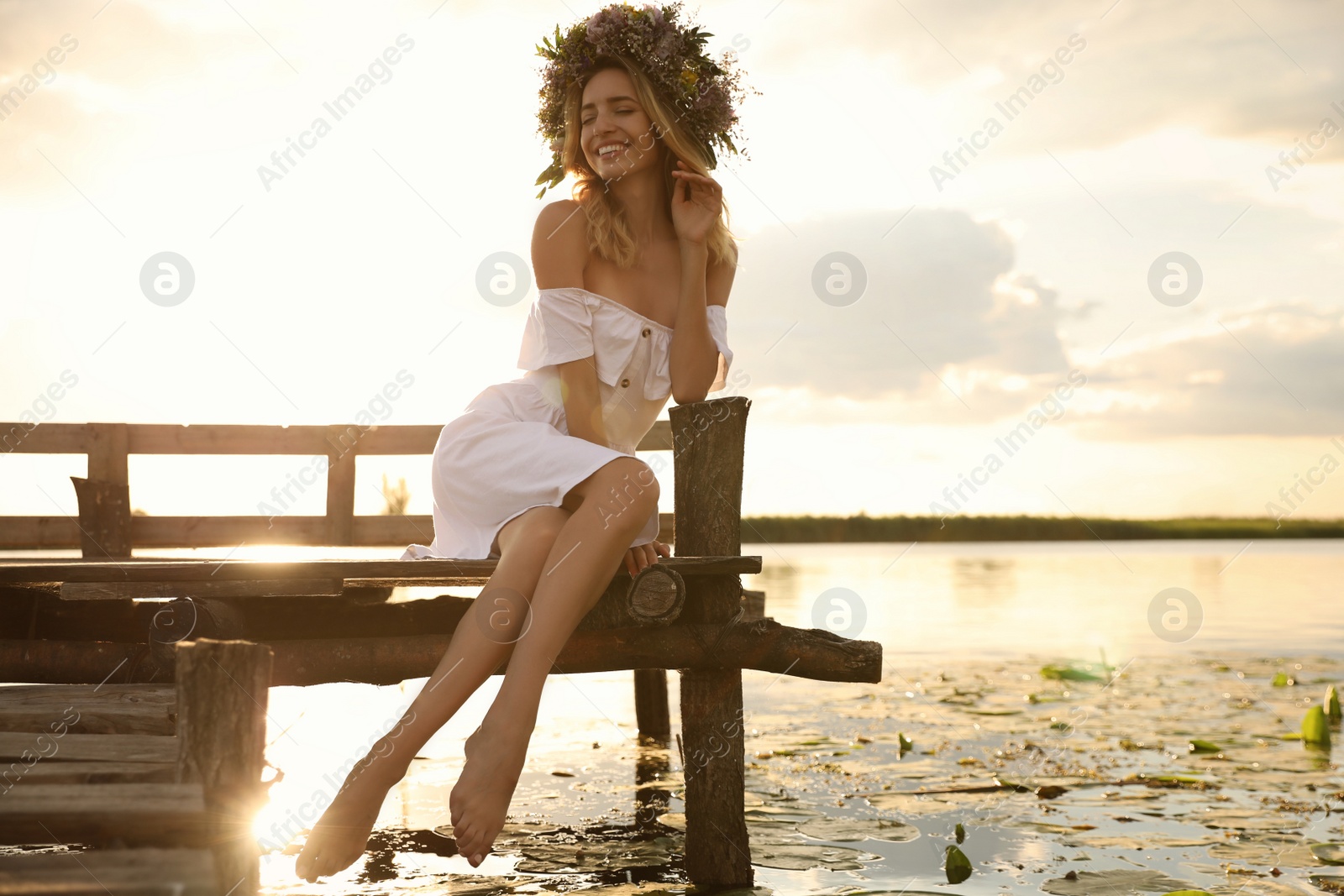 Young woman wearing wreath made of beautiful flowers on pier near river Photo of Young woman wearing wreath made of beautiful flowers on pier near river