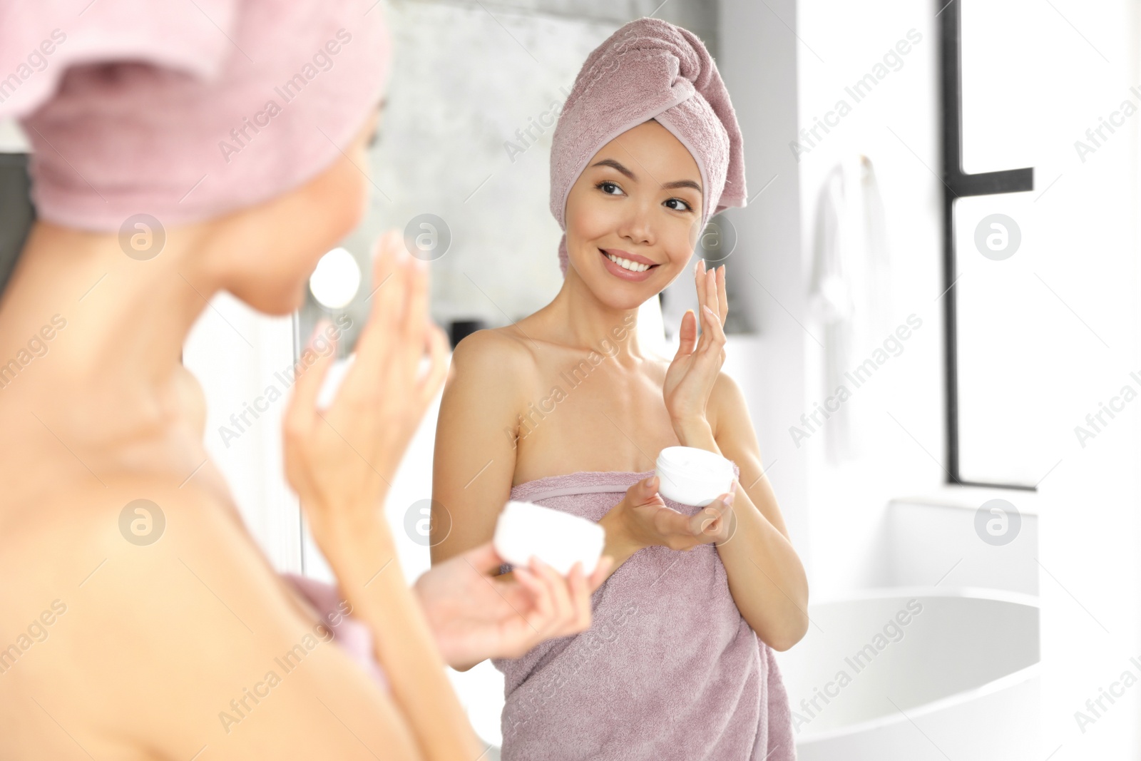 Young woman with towels applying facial cream near mirror in bathroom Photo of Young woman with towels applying facial cream near mirror in bathroom