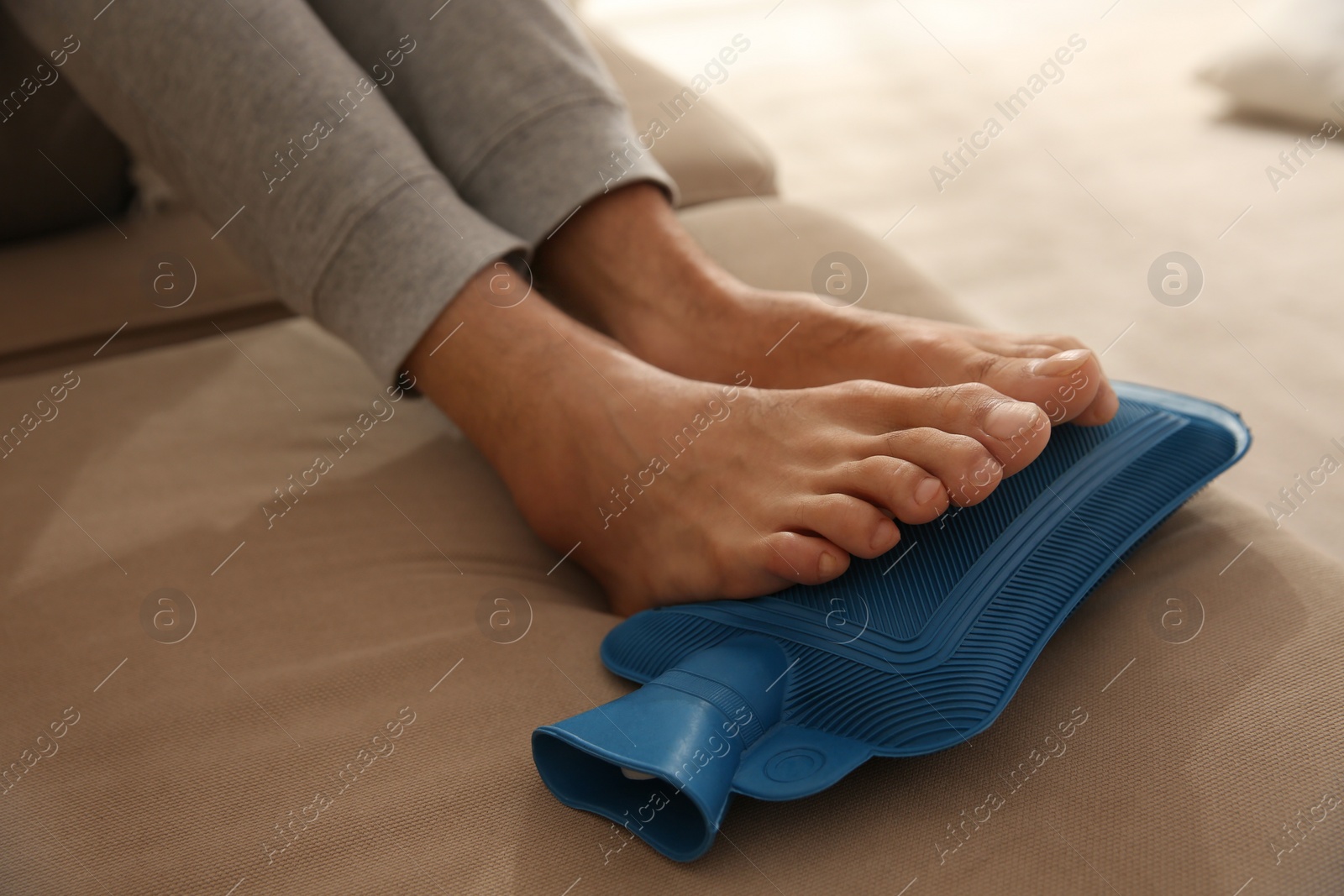Man warming feet with hot water bottle on sofa, closeup Photo of Man warming feet with hot water bottle on sofa, closeup