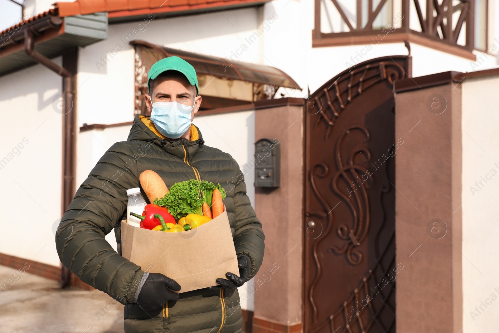 Photo of Courier in medical mask holding paper bag with groceries near house outdoors. Delivery service during quarantine due to Covid-19 outbreak