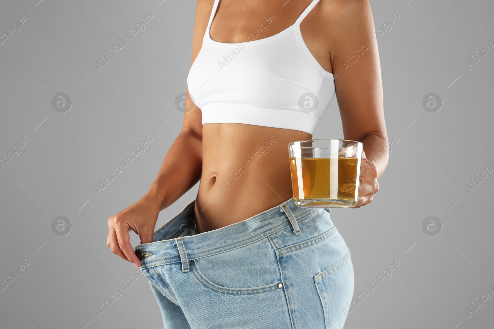 Young woman in old big jeans with cup of tea showing her diet results on grey background, closeup Photo of Young woman in old big jeans with cup of tea showing her diet results on grey background, closeup