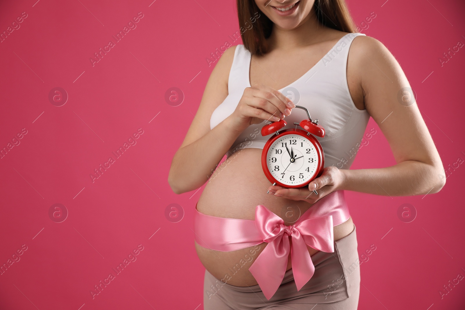 Young pregnant woman with alarm clock and bow on pink background, closeup. Time to give birth Photo of Young pregnant woman with alarm clock and bow on pink background, closeup. Time to give birth