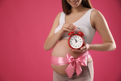Young pregnant woman with alarm clock and bow on pink background, closeup. Time to give birth Photo of Young pregnant woman with alarm clock and bow on pink background, closeup. Time to give birth
