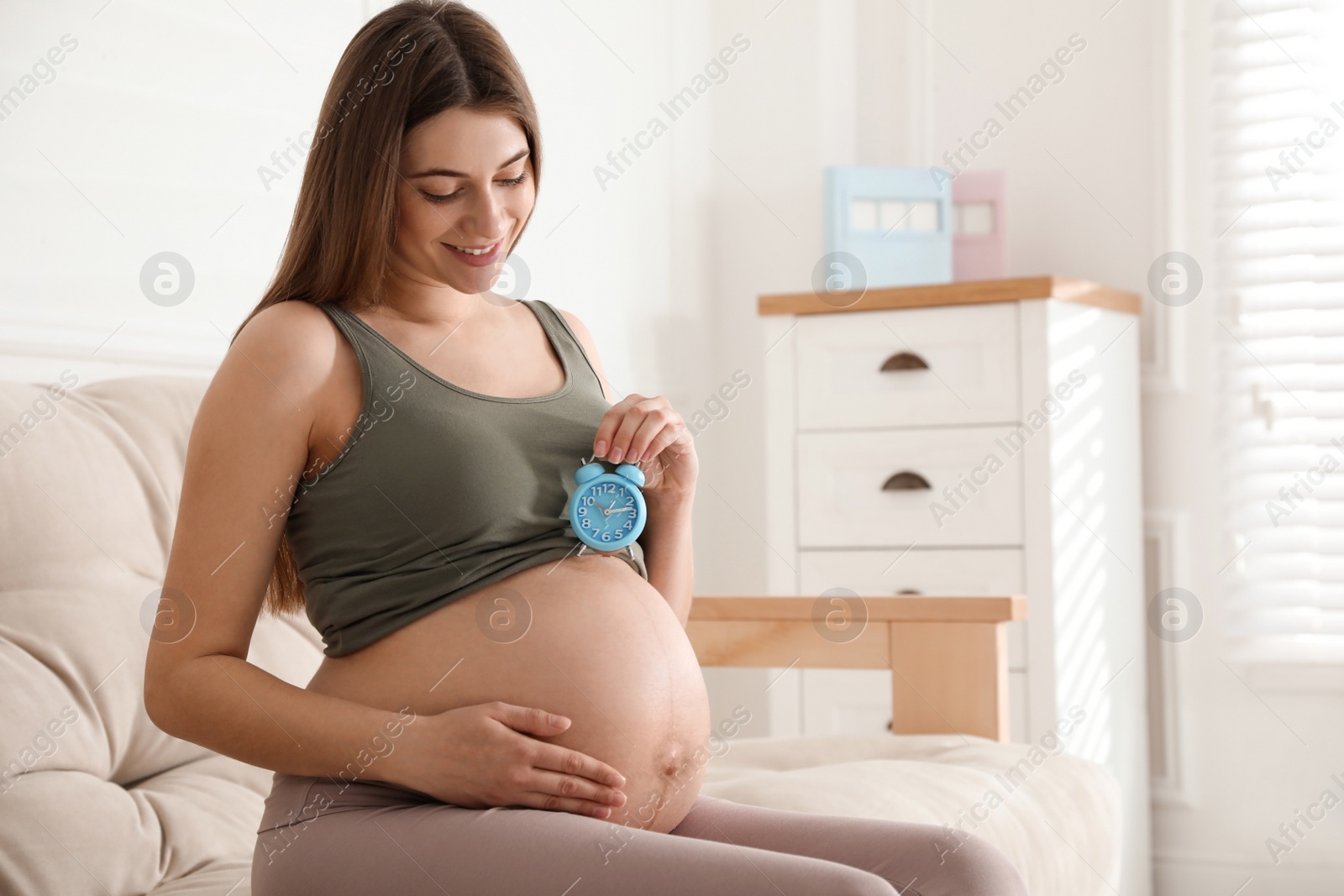 Young pregnant woman holding alarm clock near her belly at home, space for text. Time to give birth Photo of Young pregnant woman holding alarm clock near her belly at home, space for text. Time to give birth