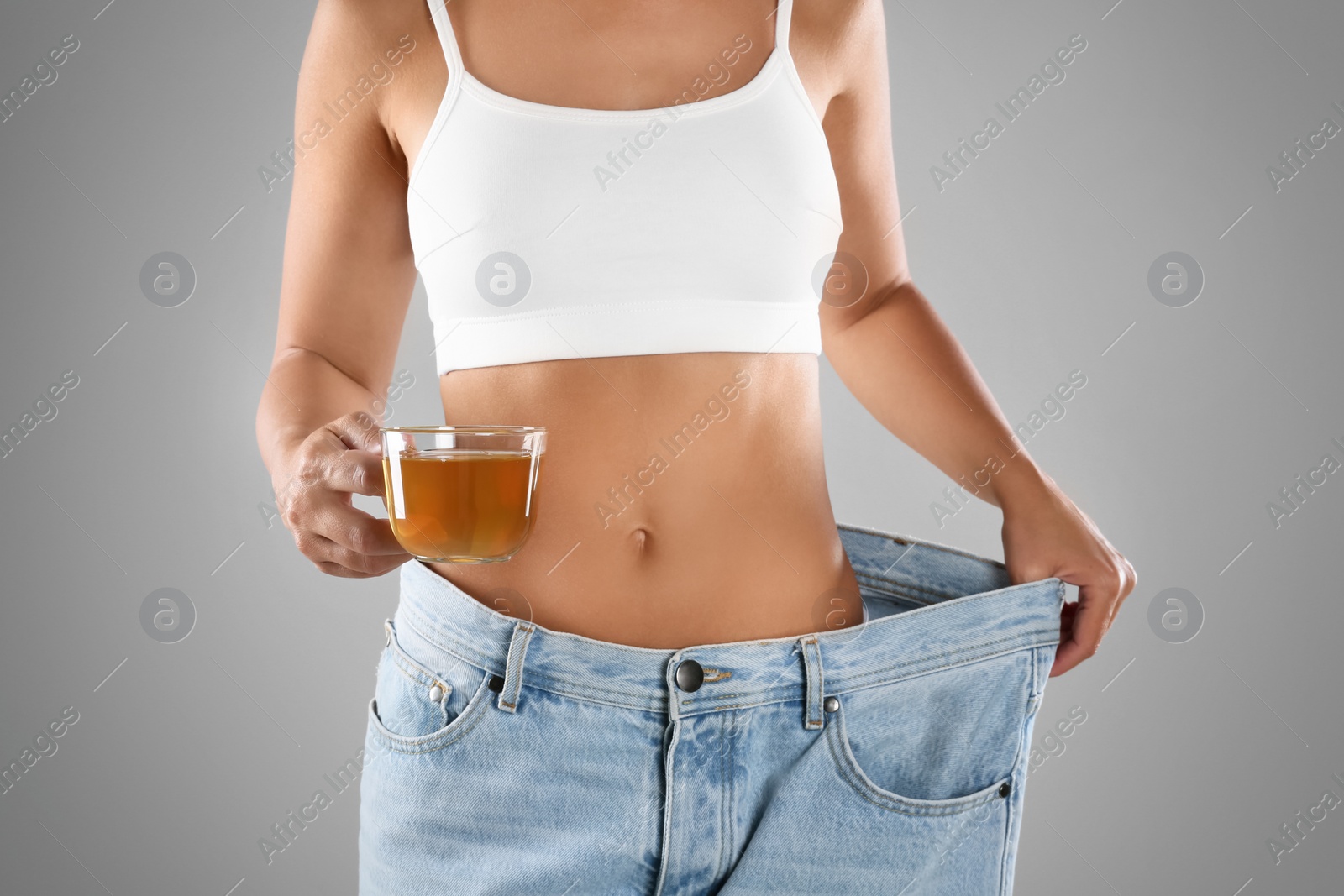Young woman in old big jeans with cup of tea showing her diet results on grey background, closeup Photo of Young woman in old big jeans with cup of tea showing her diet results on grey background, closeup