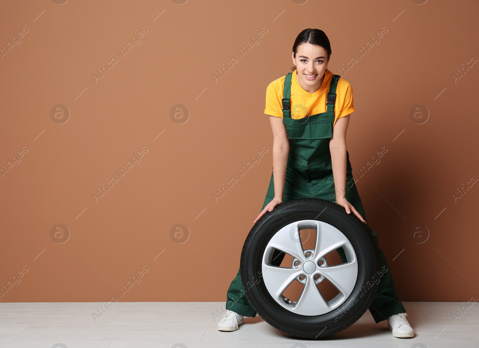 Female mechanic in uniform with car tire on color wall background Photo of Female mechanic in uniform with car tire on color wall background