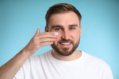Happy young man applying facial cream on light blue background Photo of Happy young man applying facial cream on light blue background