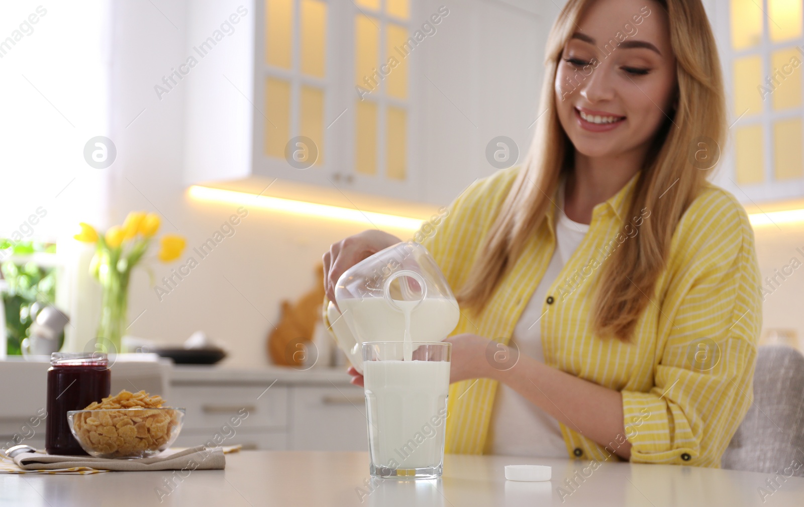 Young woman pouring milk from gallon bottle into glass at white table in kitchen Photo of Young woman pouring milk from gallon bottle into glass at white table in kitchen