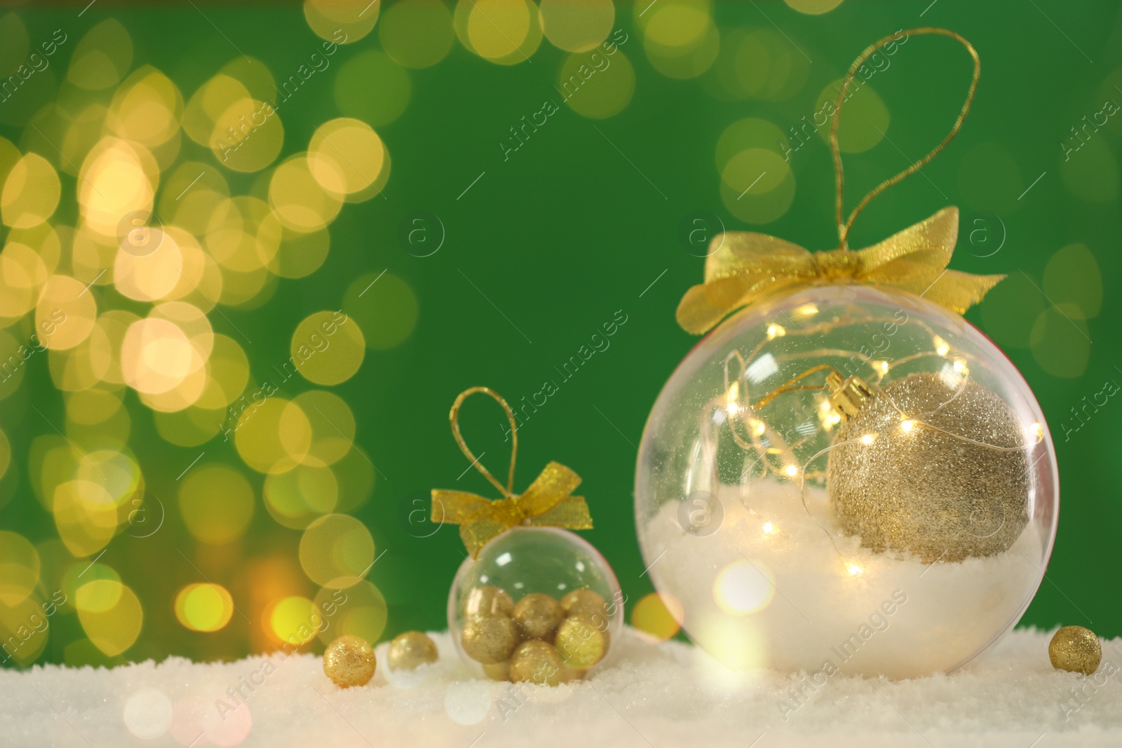 Beautiful transparent Christmas ornaments with golden bows on snow against green background, bokeh effect. Space for text Image of Beautiful transparent Christmas ornaments with golden bows on snow against green background, bokeh effect. Space for text