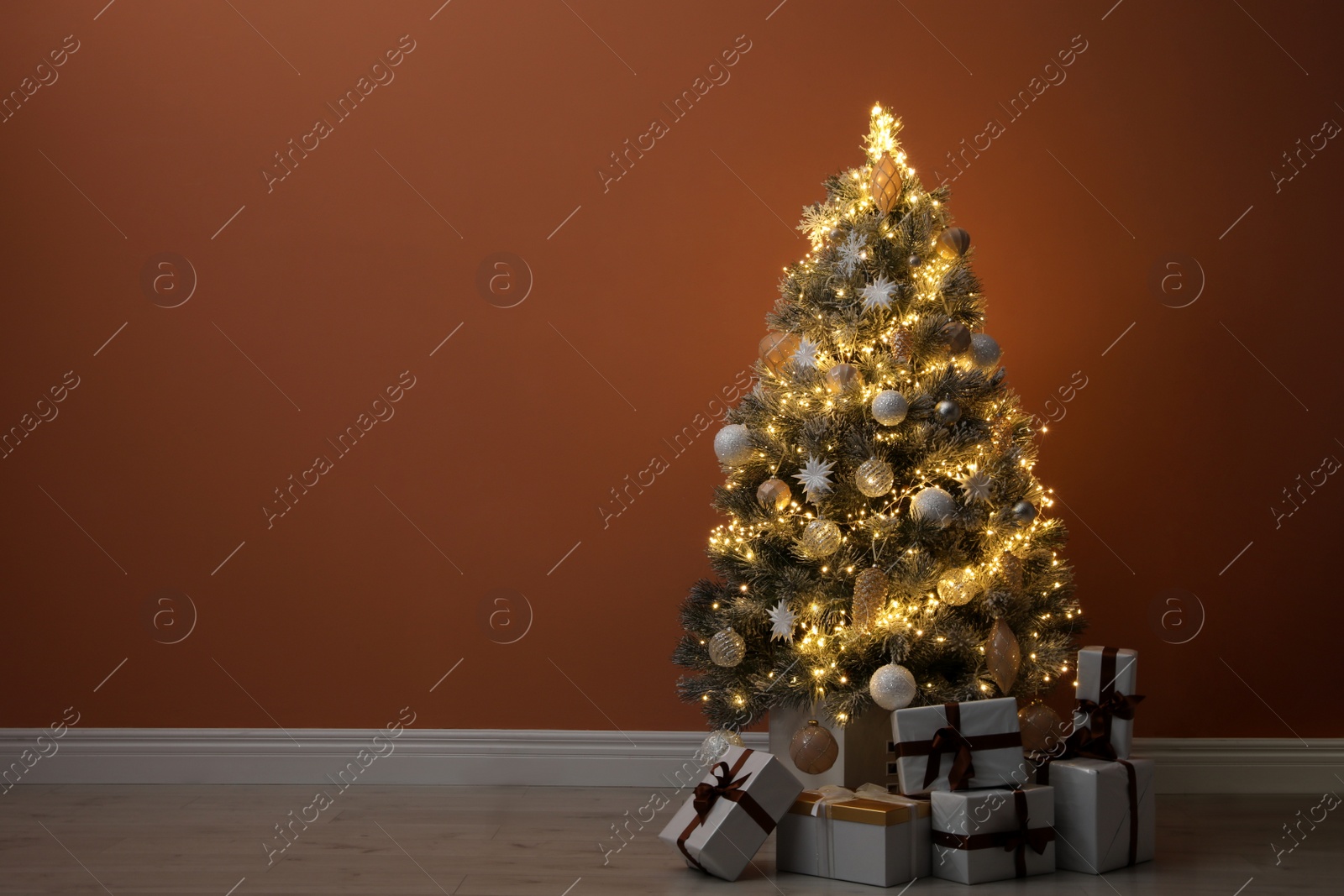 Beautiful decorated Christmas tree with glowing fairy lights and presents near orange wall indoors. Space for text Photo of Beautiful decorated Christmas tree with glowing fairy lights and presents near orange wall indoors. Space for text
