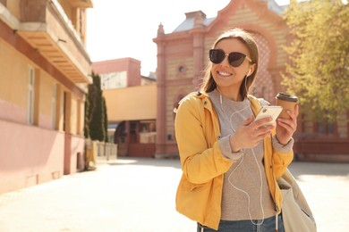 Happy young woman with coffee listening to music on city street in morning Photo of Happy young woman with coffee listening to music on city street in morning