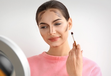 Young woman applying contouring lines on her face against light background. Professional makeup products Photo of Young woman applying contouring lines on her face against light background. Professional makeup products