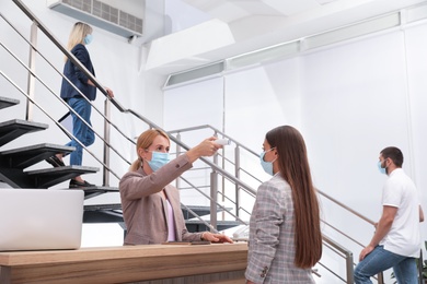 Woman in mask measuring temperature of employee with noncontact thermometer at office reception Photo of Woman in mask measuring temperature of employee with noncontact thermometer at office reception