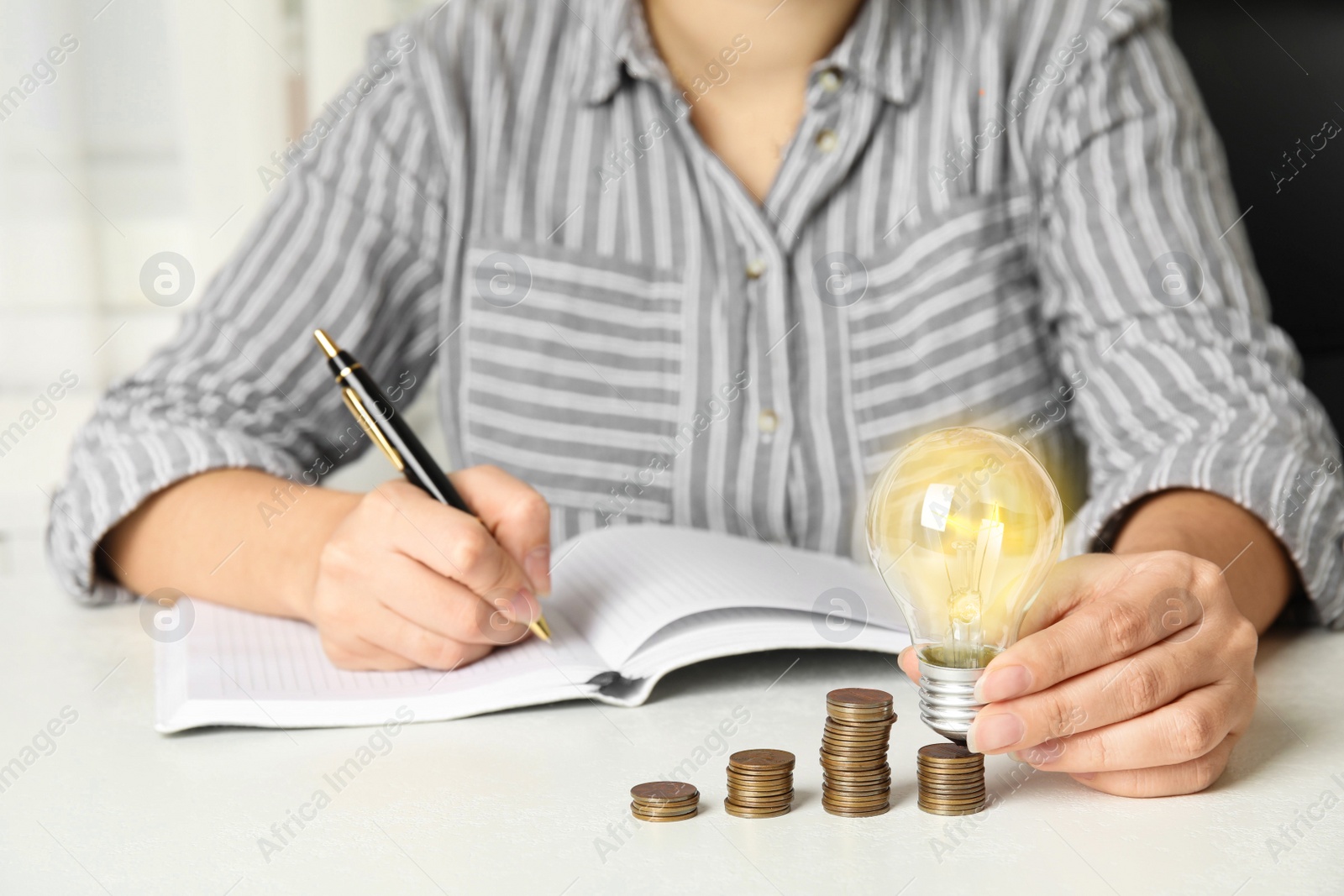 Woman with light bulb, notebook and coins at white table, closeup. Power saving Photo of Woman with light bulb, notebook and coins at white table, closeup. Power saving