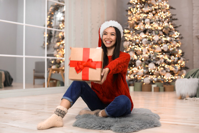 Happy young woman in Santa hat with Christmas gift at home Photo of Happy young woman in Santa hat with Christmas gift at home