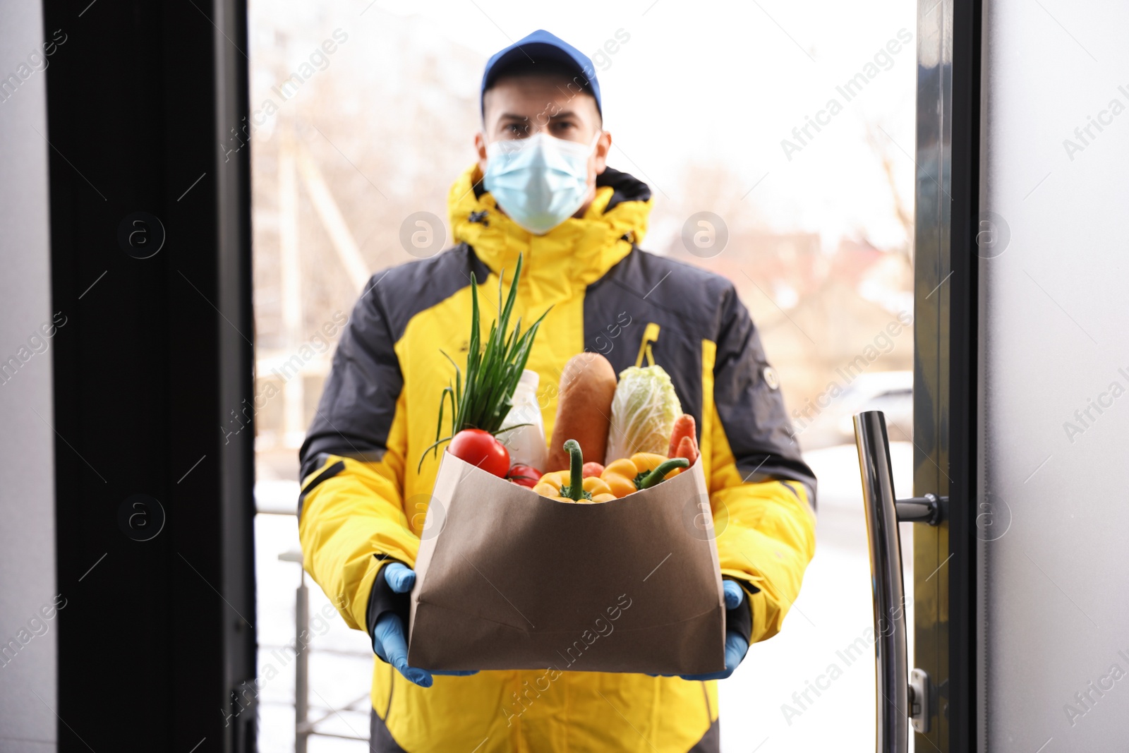 Courier in medical mask holding paper bag with groceries at doorway. Delivery service during quarantine due to Covid-19 outbreak Photo of Courier in medical mask holding paper bag with groceries at doorway. Delivery service during quarantine due to Covid-19 outbreak