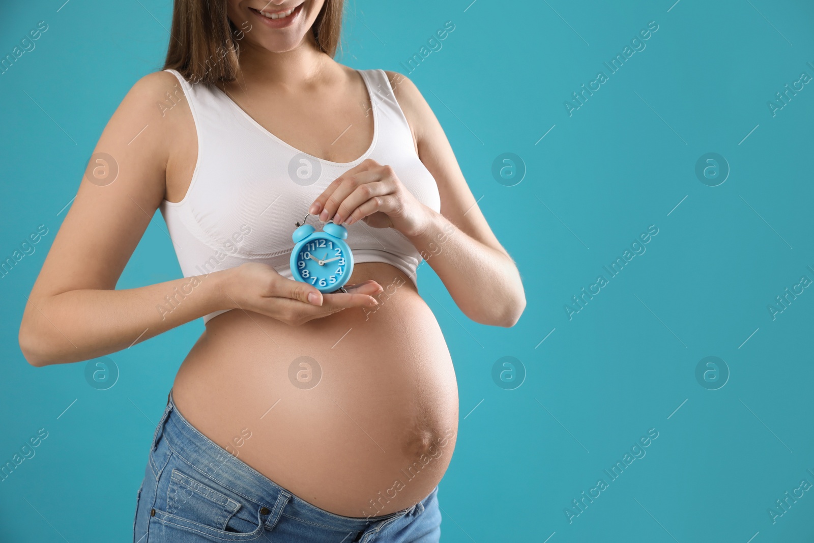 Young pregnant woman holding alarm clock near her belly on light blue background, closeup and space for text. Time to give birth Photo of Young pregnant woman holding alarm clock near her belly on light blue background, closeup and space for text. Time to give birth