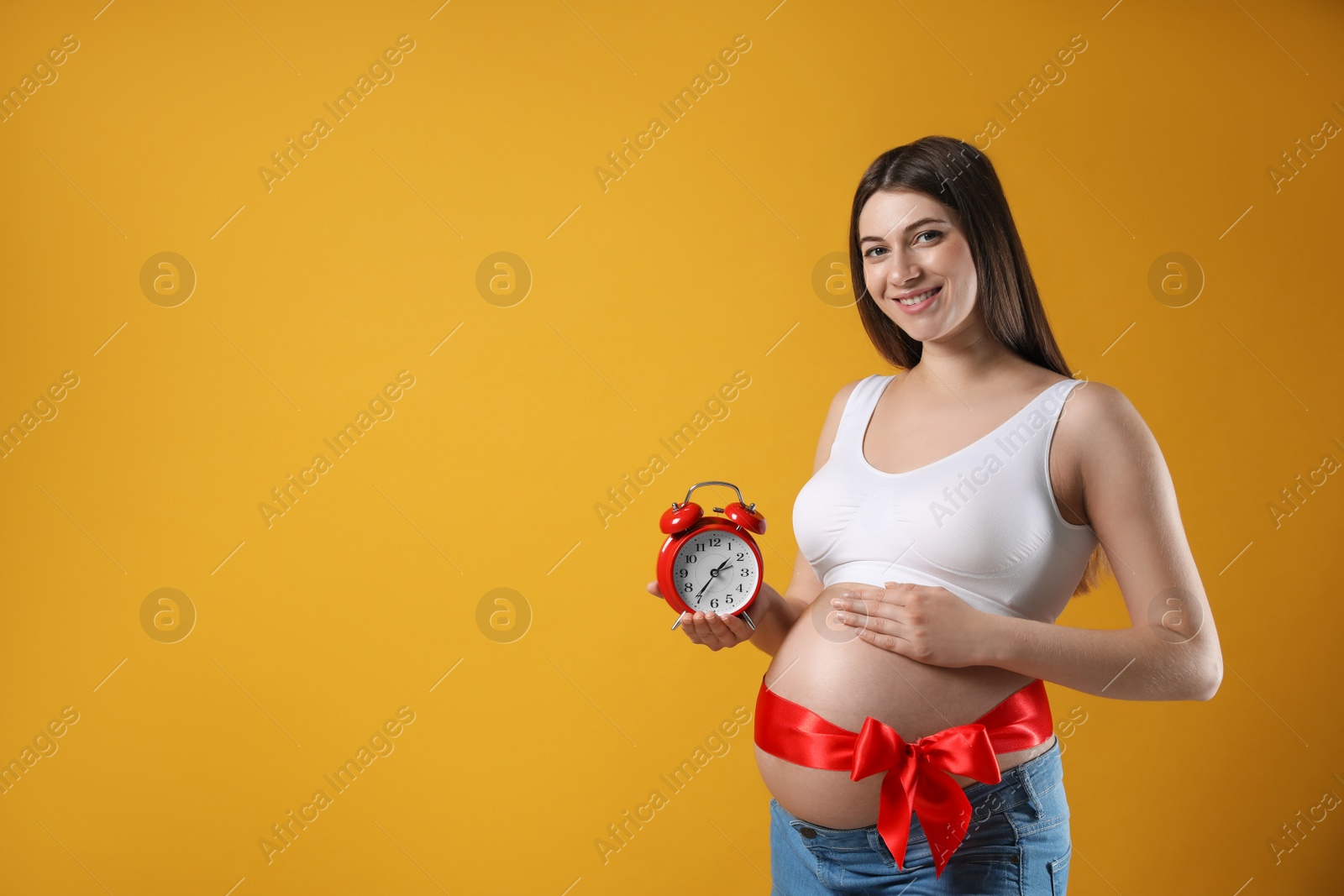 Young pregnant woman with alarm clock and red bow on yellow background, space for text. Time to give birth Photo of Young pregnant woman with alarm clock and red bow on yellow background, space for text. Time to give birth